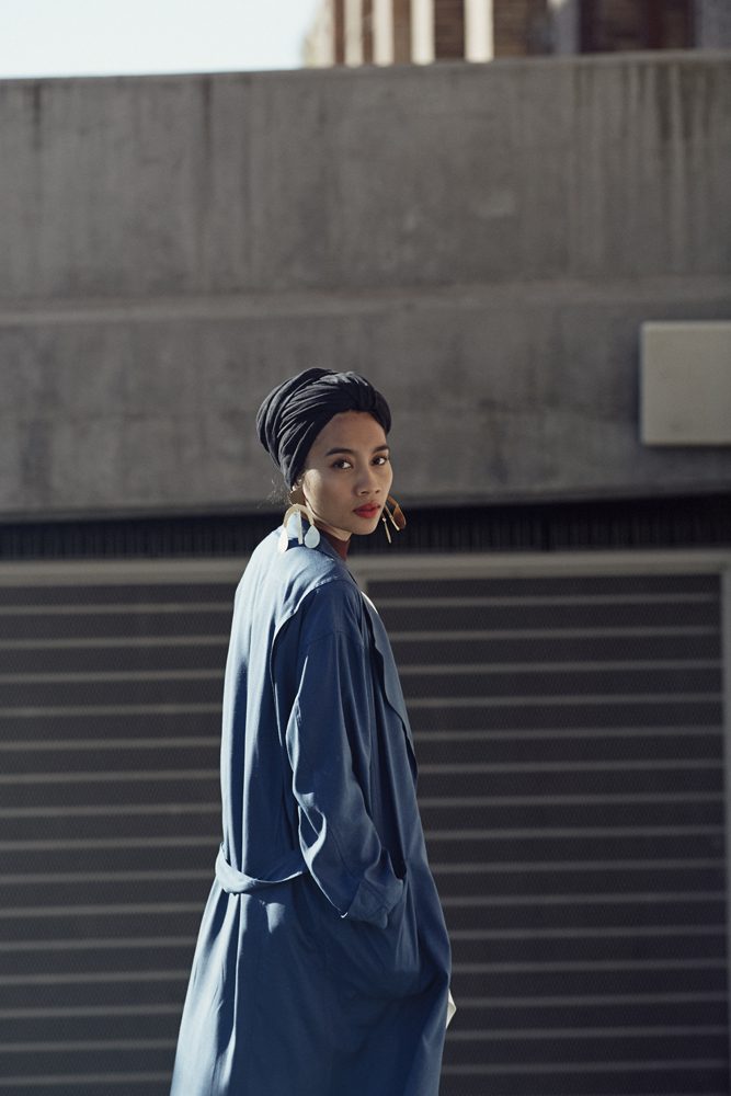 A woman in a long blue coat and dark head wrap standing beside a concrete wall.