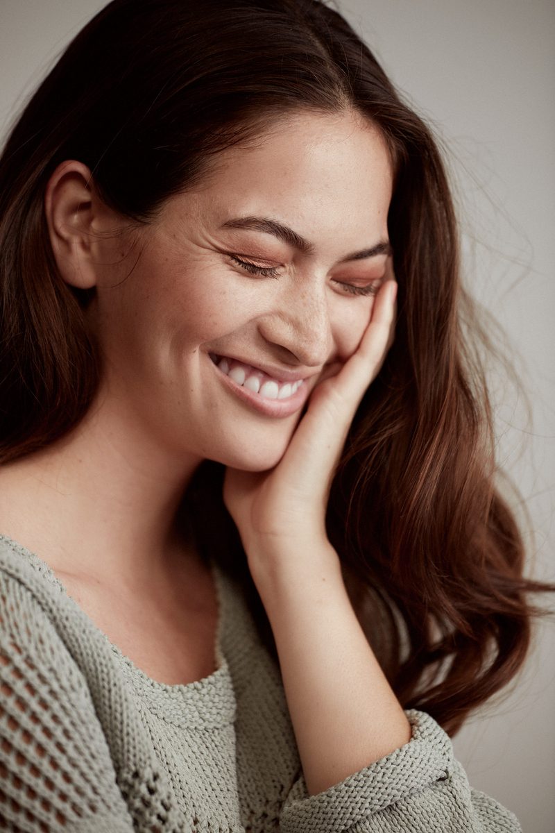 A woman with long brown hair smiling with her eyes closed and her hand resting against her cheek.