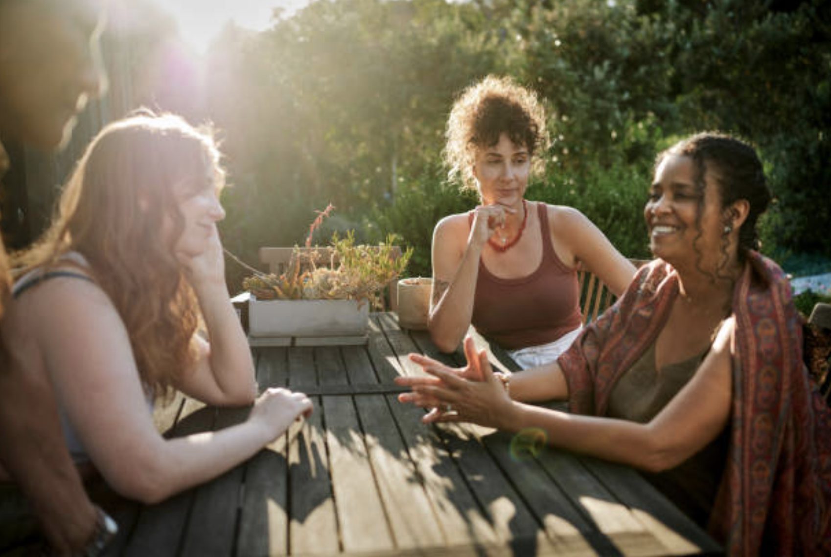Three women talking at an outdoor wooden table in late afternoon sun.